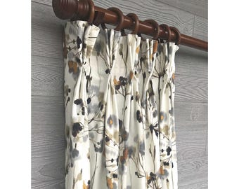 May include: A white curtain panel featuring a printed pattern of abstract dandelion seed heads in shades of gray, black, and muted gold. The fabric is hung from a dark brown wooden curtain rod with matching rings, creating deep vertical pleats.