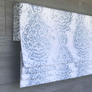 Puede incluir: Una cortina romana con un estampado de damasco blanco y azul. La tela está doblada en pliegues horizontales. La cortina está montada sobre un marco de madera. El patrón es un diseño floral estilizado a gran escala.