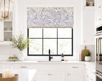 May include: A fabric roman shade featuring a grey and white paisley pattern installed over a black-framed window. The shade is mounted inside the window frame and hangs above a white kitchen sink with a black faucet and white cabinetry.