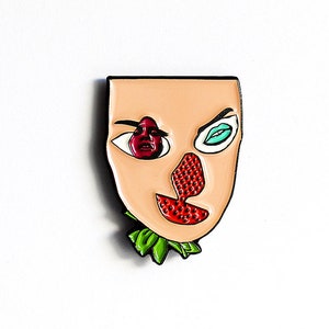 May include: A colorful enamel pin featuring a stylized face with a strawberry nose, a red eye, and a blue eye. The pin is on a white card with the text "blond eyed girl" in a black diamond shape.