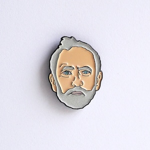 Puede incluir: Un pin con un retrato estilizado de un hombre con cabello gris y barba, sobre un fondo blanco. El pin tiene una cara marrón claro con ojos azules. Las palabras "blond eyed girl" están impresas en la parte inferior de la tarjeta.