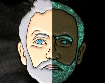 以下が含まれることがあります： 白とグレーのエナメル製のピンバッジで、男性の顔とひげが描かれています。ピンバッジは暗闇で光り、ひげと目は緑色に光ります。