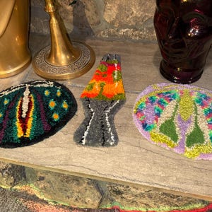 May include: Three colorful, hand-stitched rug designs. One is a green and purple butterfly, another is a multi-colored lava lamp, and the last is a purple and green butterfly.
