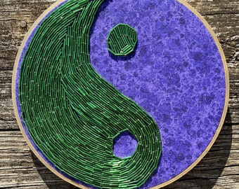 Puede incluir: Un símbolo yin yang de cuentas verdes y moradas sobre un fondo de madera. El símbolo está hecho de pequeñas cuentas verdes y está rodeado de un fondo morado.
