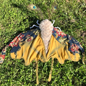 May include: A yellow, gold, and blue butterfly costume with floral fabric accents. The costume has a furry body and long, flowing wings.