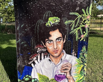 Puede incluir: Una manta de franela negra y verde con una ilustración colorida de una persona sosteniendo un vaso de líquido morado. La persona está rodeada de plantas, hongos y una rana verde.