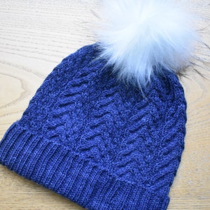 May include: A blue knitted beanie with a white pom pom. The beanie has a cable knit pattern.