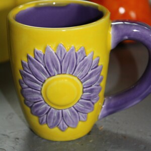 Pode incluir: Uma caneca de cerâmica amarela com um desenho de flor roxa. A caneca tem uma alça roxa.
