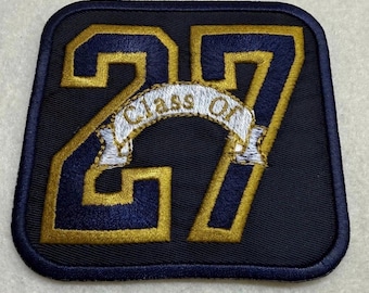Könnte beinhalten: Ein quadratischer gestickter Aufnäher mit abgerundeten Ecken, der die großen Zahlen 27 in Goldfaden mit dunkelblauem Rand zeigt. Ein weißes Banner ist mittig aufgestickt mit dem Text Class Of in Goldfaden.
