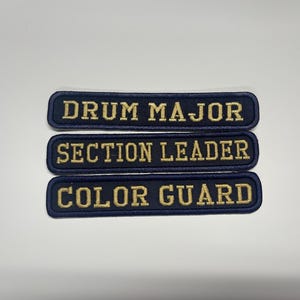 May include: Three rectangular embroidered patches with dark blue fabric and gold lettering. The top patch reads DRUM MAJOR, the middle patch reads SECTION LEADER, and the bottom patch reads COLOR GUARD. Each patch features a dark blue stitched border.