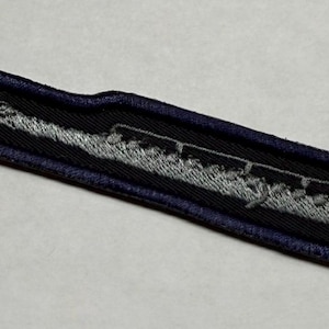May include: A long, narrow, rectangular embroidered patch featuring a silver-gray flute design centered on a black background. The patch is finished with a dark navy blue stitched border that follows the elongated shape of the musical instrument.