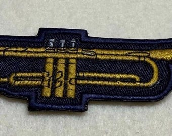 Könnte beinhalten: Ein gestickter Aufnäher in Form einer Trompete, der goldenes Garn für den Instrumentenkörper und den Schallbecher sowie silbernes Garn für das Mundstück und die Ventile verwendet, alles vor einem dunkelblauen Hintergrund mit passendem Rand.