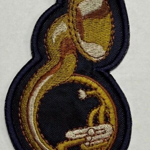 May include: An embroidered patch featuring a gold and brown sousaphone musical instrument. The design is outlined with a dark navy blue border and filled with detailed stitching in shades of yellow, gold, brown, and white to depict the brass instrument.