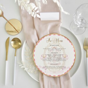 May include: A formal table setting with a white plate, gold utensils, and a menu card. The menu card is round with a pink and gold design and lists starters, entree, and dessert options.