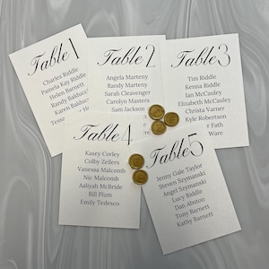 May include: Five white place cards with black calligraphy, labeled "Table 1" through "Table 5", listing names. Gold wax seals with the letter "E" are scattered around the cards. The background is a light, marbled pattern.