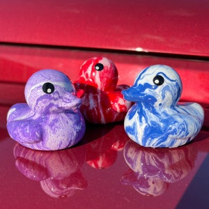May include: Three decorative duck figurines in purple, red, and blue with white marbling. Each duck has a black eye and a beak. The ducks are arranged in a row, with the purple duck on the left, the red in the middle, and the blue on the right.
