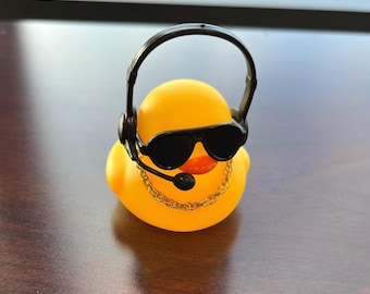 May include: A yellow rubber duck wearing black sunglasses, a headset, and a gold chain. The duck has an orange beak and is a fun, novelty item.
