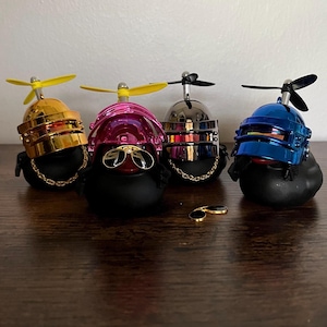 May include: Four black duck figurines with metallic helmets in gold, pink, silver, and blue, each with a propeller. One pair of gold sunglasses rests nearby. The ducks have gold chain necklaces.