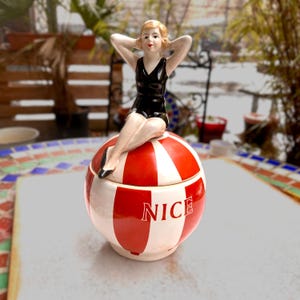 Könnte beinhalten: Ein Vintage-Keramiktopf mit Deckel, mit einem rot-weiß gestreiften Design. Eine Figur einer Frau in einem schwarzen Badeanzug sitzt oben. Das Wort "NICE" ist auf dem Topf abgebildet.