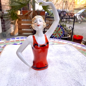 Könnte beinhalten: Porzellanfigur einer Frau in einem roten Badeanzug mit schwarzen Streifen, ein Arm erhoben. Die Figur hat blondes Haar und posiert in einer anmutigen, künstlerischen Haltung. Die Figur steht auf einer weißen Oberfläche.