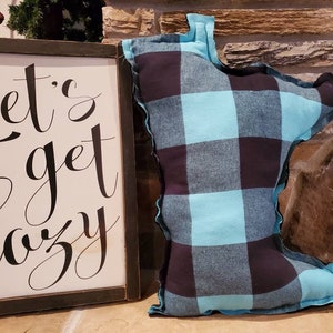May include: A blue and black plaid pillow shaped like the state of Minnesota. The pillow is sitting on a stone surface next to a wooden sign that says "Let's get cozy".