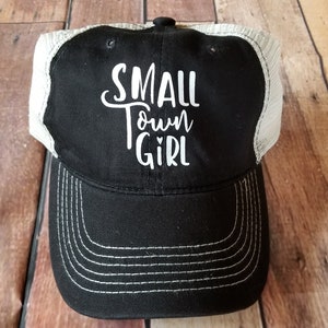 May include: Black and white trucker hat with a white embroidered design that says "SMALL town GIRL" with a small heart in the word "town".