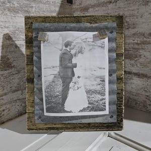 May include: A rustic wooden picture frame with a corrugated metal border. The frame holds a black and white photograph of a couple kissing. The frame is secured with small wooden blocks and nails, creating a vintage aesthetic.