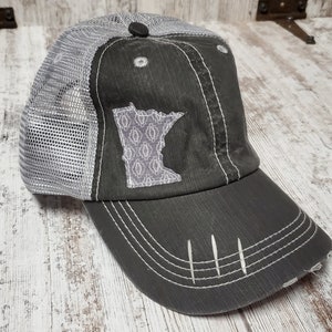 May include: A gray and white mesh trucker hat with a distressed look. The hat features a gray outline of the state of Minnesota with a white patterned design inside.
