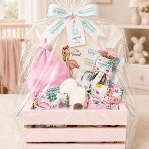 May include: A baby gift set in a light pink wooden crate wrapped in clear cellophane. It includes a cream-colored onesie with a flamingo graphic and the text I've Arrived!, a pink hat, a white cloth, and a sensory teether with a flamingo print.