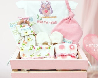 May include: A pink wooden crate filled with baby items. Includes a white onesie with an owl graphic, a pink hat, mittens, a wooden teether, and floral print items. The text "Moo's the cutest!" is on the onesie.