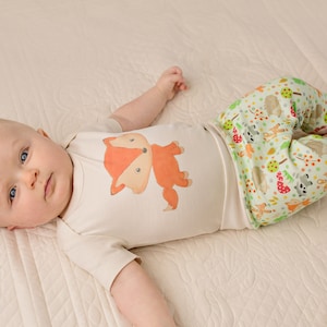 May include: A baby wearing a white onesie with a fox graphic and green pants with a woodland animal print.