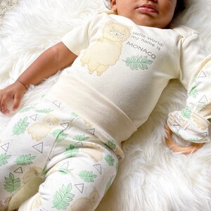 May include: A baby wearing a cream colored onesie with a cartoon llama and the text "Hello World! My name is MONACO". The baby is also wearing white and green pants with a llama and leaf pattern.