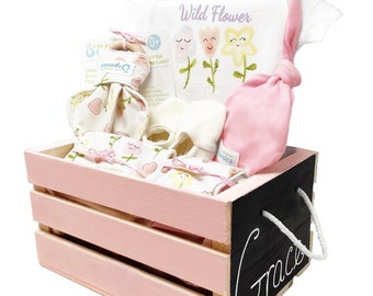 May include: A pink wooden crate with a black chalkboard side that says "Grace". The crate is filled with baby items, including a white onesie with a flower design, a pink hat, and a pair of white booties.