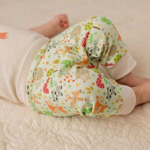 May include: A baby wearing a white onesie with a fox print and green and white pants with a woodland animal print.