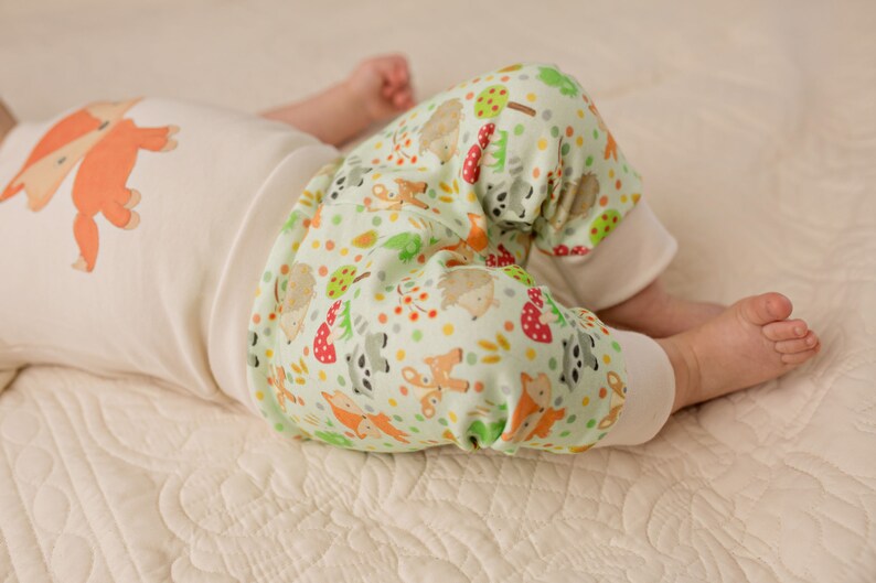 May include: A baby wearing a white onesie with a fox print and green and white pants with a woodland animal print.