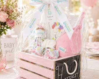 May include: A pink wooden crate gift set wrapped in clear cellophane with a white ribbon printed with I've Arrived Organics. The set includes a pink knotted baby hat, a white onesie with a unicorn graphic, and patterned baby items.