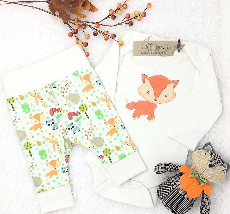 May include: A white baby onesie with long sleeves features a cartoon fox illustration and the text "I've Arrived Organics". A pair of white baby leggings with a green and white woodland animal print are also pictured. A brown and black stuffed animal fox is included.