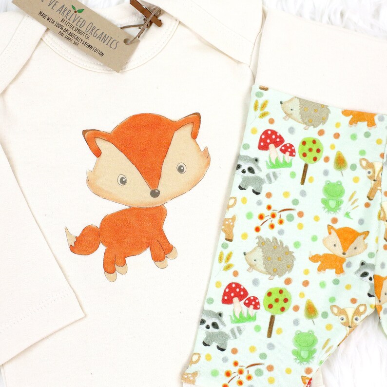 May include: A white baby onesie with a cute orange fox illustration. The onesie is paired with green and white leggings featuring a woodland pattern with foxes, raccoons, hedgehogs, mushrooms, and trees.