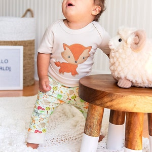 May include: A baby wearing a cream colored shirt with a fox graphic and green and white patterned pants stands on a wooden stool. A white plush lamb sits on the stool. The sign behind the baby reads "Hello World!"