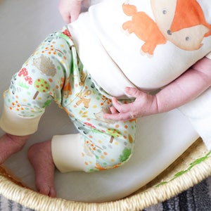 May include: A baby wearing a white onesie with a fox graphic and green and white pants with a woodland animal print. The baby is lying in a woven basket.