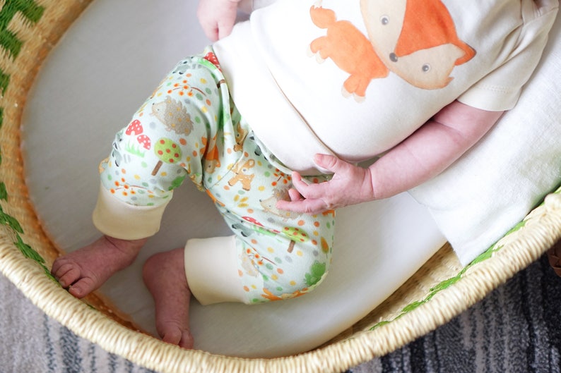 May include: A baby wearing a white onesie with a fox graphic and green and white pants with a woodland animal print. The baby is lying in a woven basket.