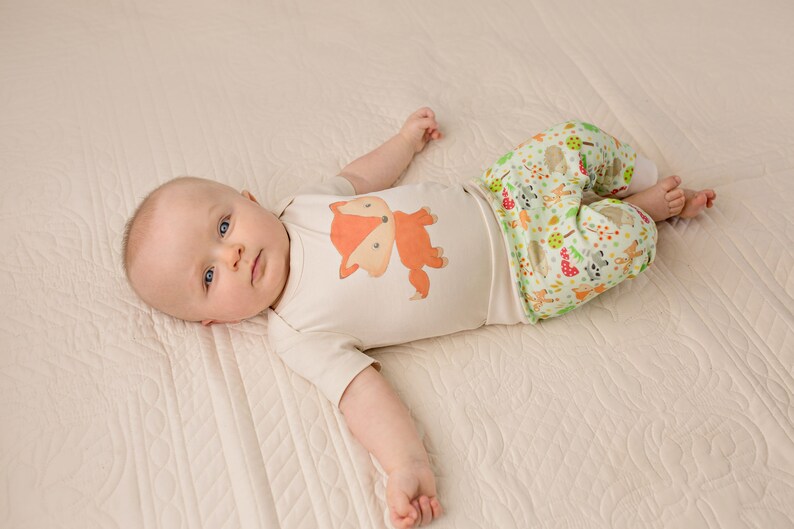 May include: A baby wearing a white onesie with a fox graphic and green pants with a woodland animal print.