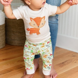 May include: A baby wearing a white bodysuit with a cartoon fox print and green pants with a woodland animal print.