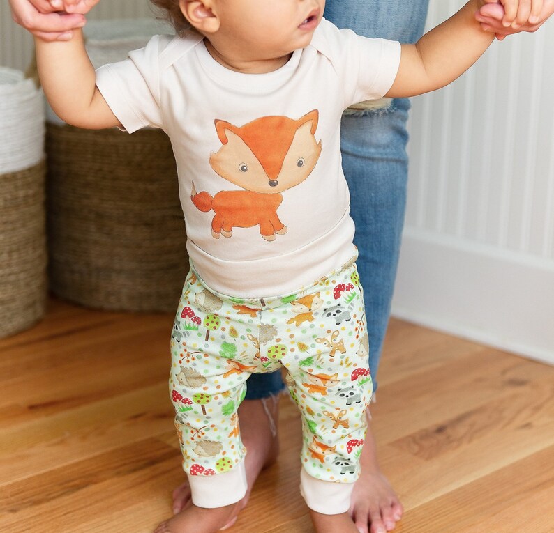 May include: A baby wearing a white bodysuit with a cartoon fox print and green pants with a woodland animal print.
