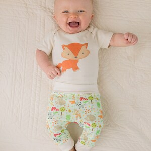 May include: A baby wearing a white bodysuit with a fox print and green pants with a woodland animal print. The baby is lying on a white blanket and smiling.