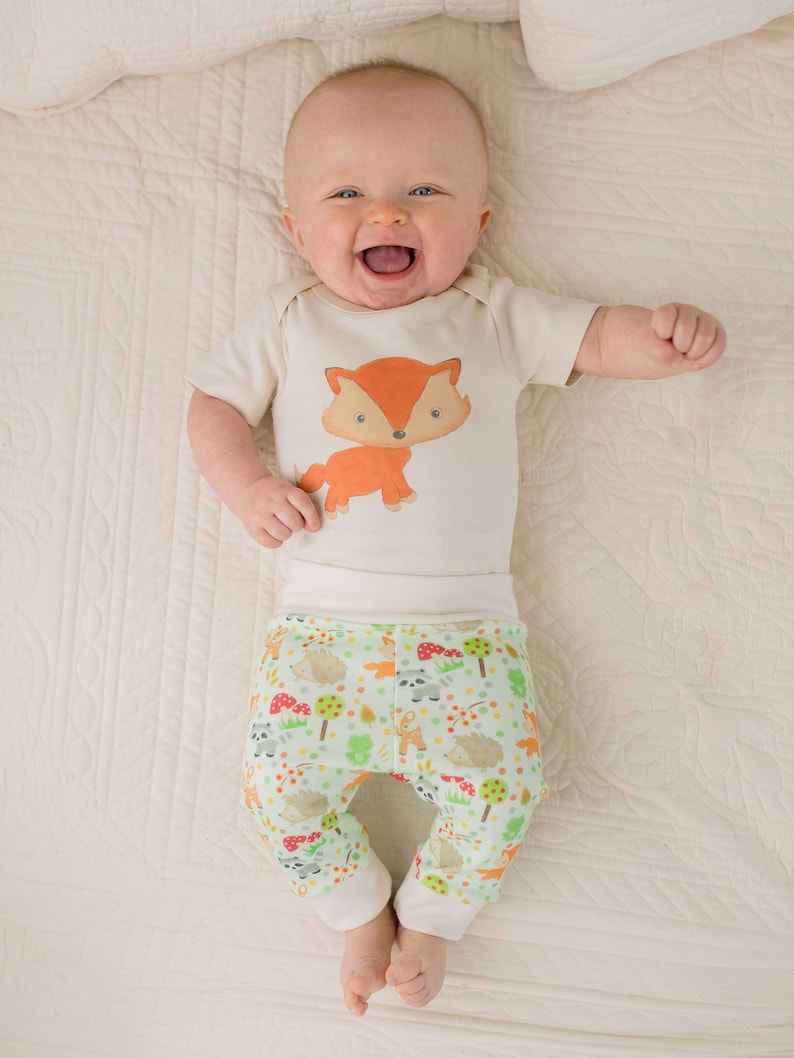 May include: A baby wearing a white bodysuit with a fox print and green pants with a woodland animal print. The baby is lying on a white blanket and smiling.