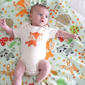 May include: A baby wearing a white onesie with a fox print on a green and white blanket with a woodland animal print. The blanket features foxes, deer, raccoons, hedgehogs, mushrooms, and leaves.