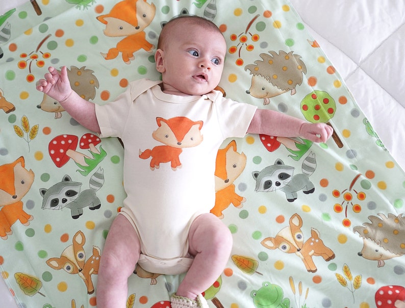 May include: A baby wearing a white onesie with a fox print on a green and white blanket with a woodland animal print. The blanket features foxes, deer, raccoons, hedgehogs, mushrooms, and leaves.
