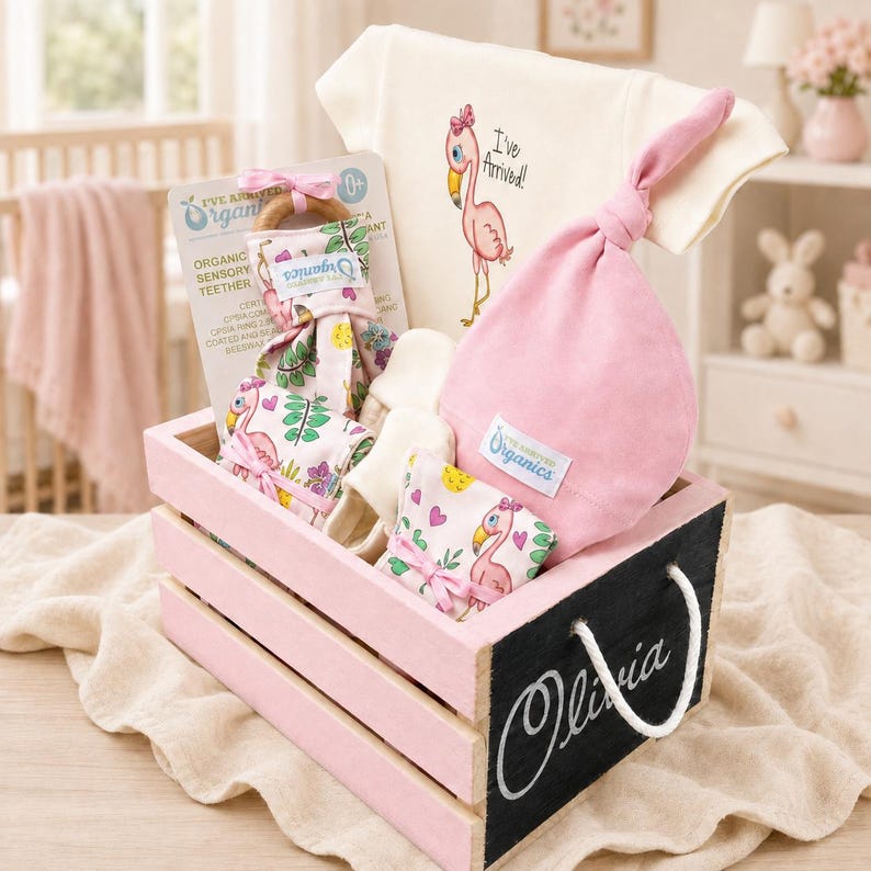 May include: A baby gift set in a pink wooden crate labeled Olivia, containing a cream onesie with a flamingo graphic and text I've Arrived, a pink knotted baby hat, a wooden teether with a floral flamingo fabric, and cream-colored baby booties.