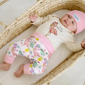 May include: A baby outfit consisting of a cream-colored long-sleeve shirt with a pink flamingo graphic and the text "I've Arrived!", pink pants with a flamingo and floral pattern, and a matching pink knotted beanie hat with a small white label.